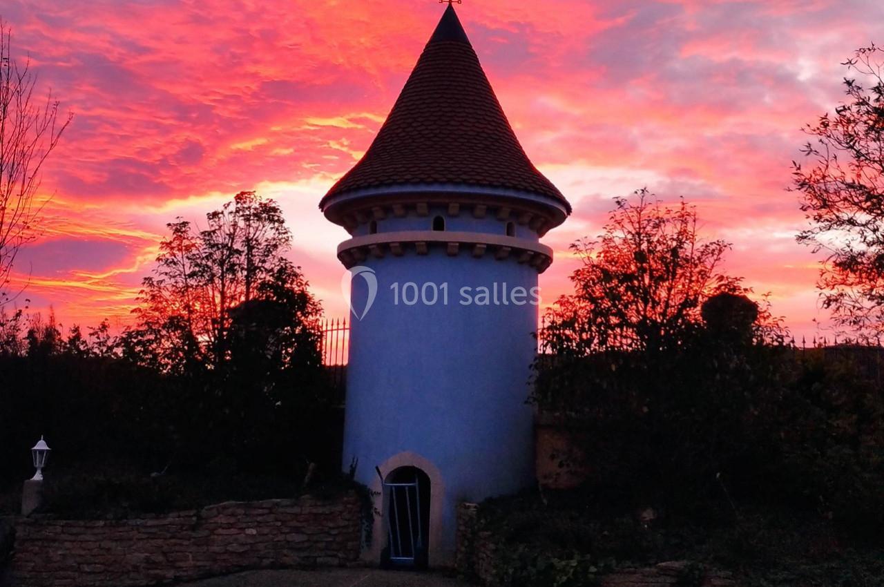 Tour ronde bleue avec un toit en tuiles rouges, entourée de végétation, sous un ciel rougeoyant au coucher du soleil.