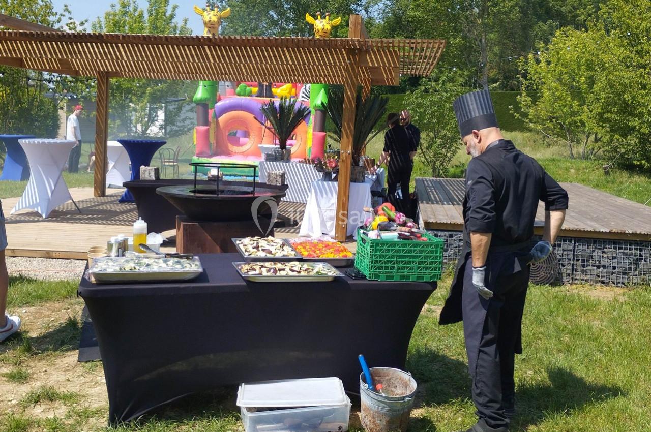 Un chef en tenue noire prépare un buffet en plein air avec des plats disposés sur une table, dans un cadre verdoyant.