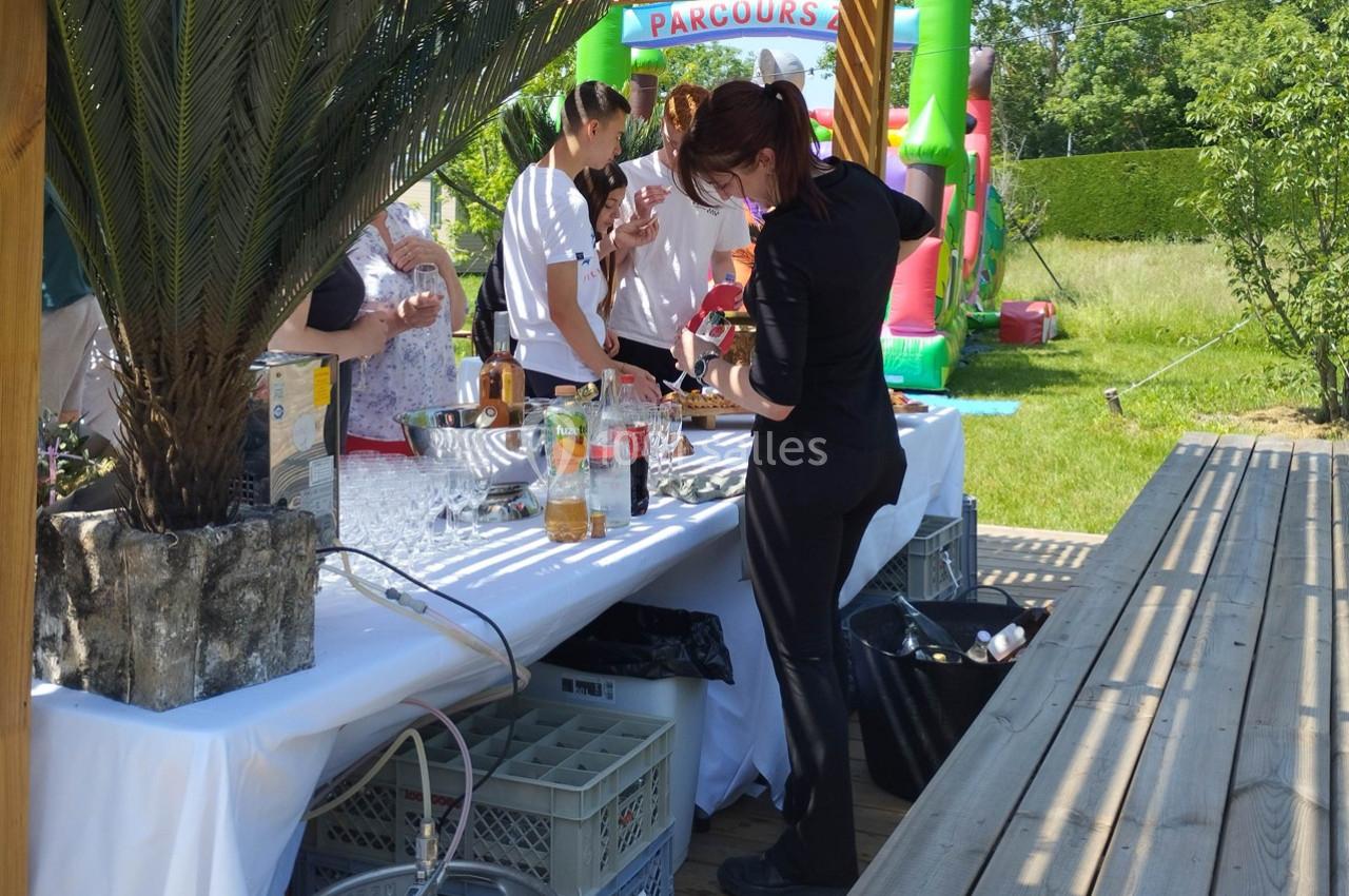 Une femme prépare des boissons sur une table de buffet en extérieur, avec des invités et un château gonflable en arrière…