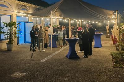 Espace extérieur couvert par une tente beige, avec des tables et des personnes assises dans un cadre verdoyant.