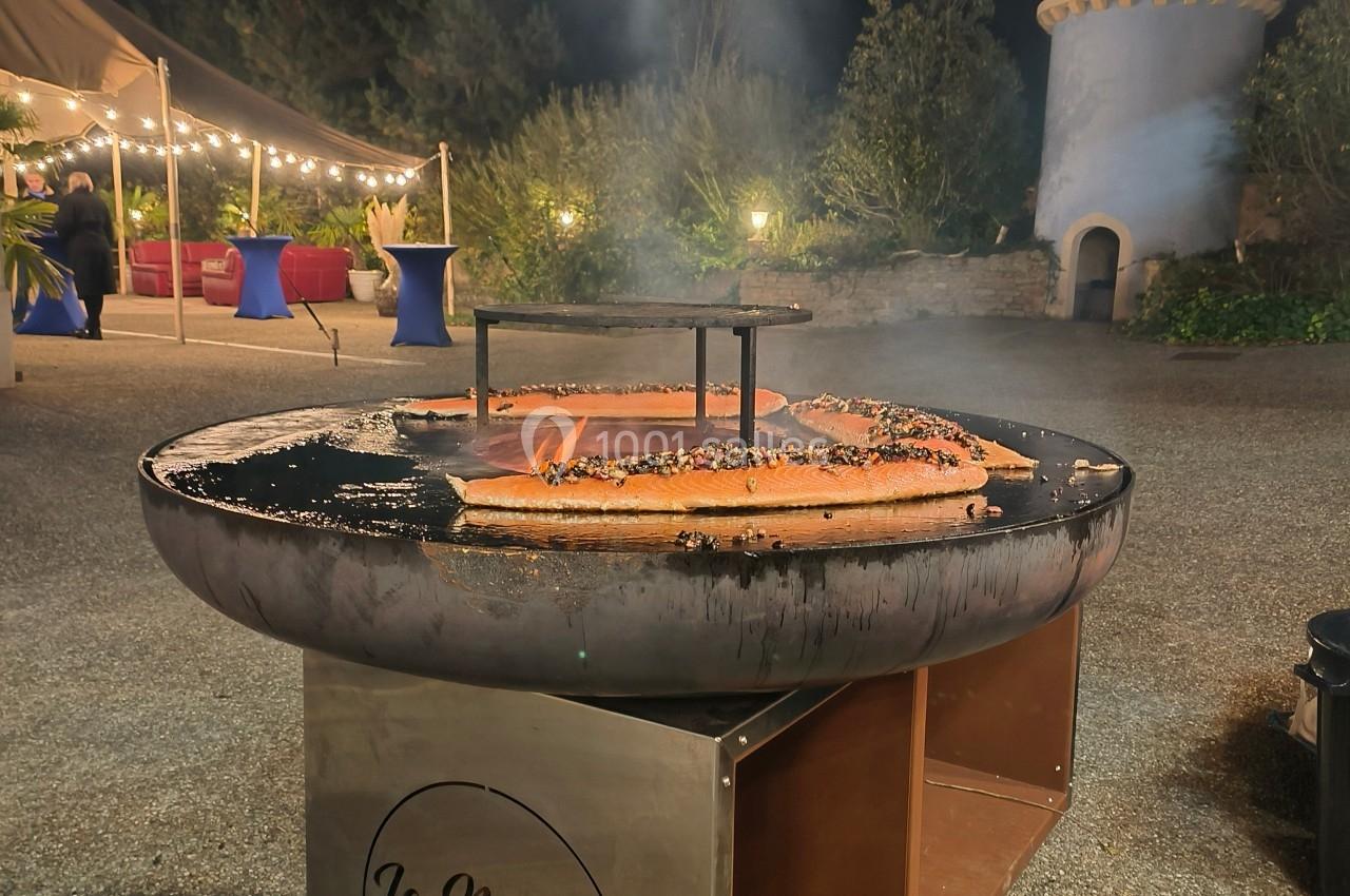 Barbecue circulaire en extérieur, avec des filets de saumon en cuisson, devant un décor nocturne et une tour en arrière-plan.
