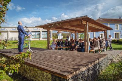 Espace extérieur couvert par une tente beige, avec des tables et des personnes assises dans un cadre verdoyant.