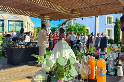 Espace extérieur couvert par une tente beige, avec des tables et des personnes assises dans un cadre verdoyant.