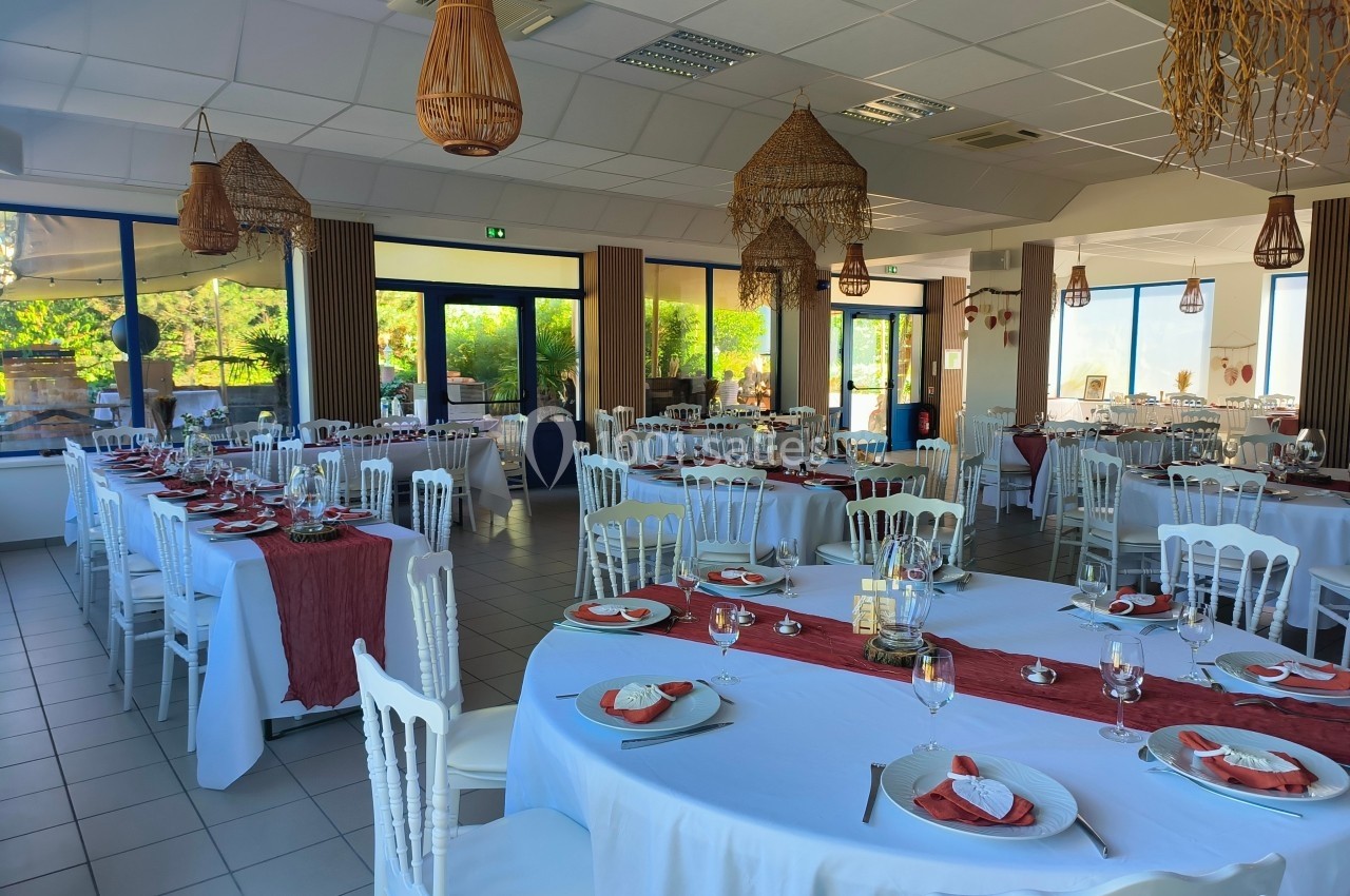 Salle de réception lumineuse avec tables dressées, nappes blanches et décorations rouges, ambiance élégante et conviviale.