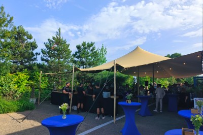 Espace extérieur couvert par une tente beige, avec des tables et des personnes assises dans un cadre verdoyant.