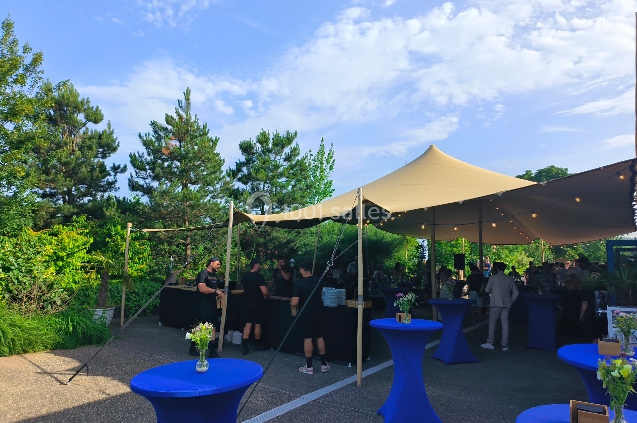 Tente beige montée en extérieur avec des tables hautes bleues et des personnes rassemblées près d'un bar.