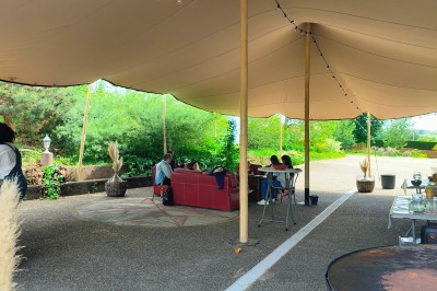 Espace extérieur couvert par une tente beige, avec des tables et des personnes assises dans un cadre verdoyant.