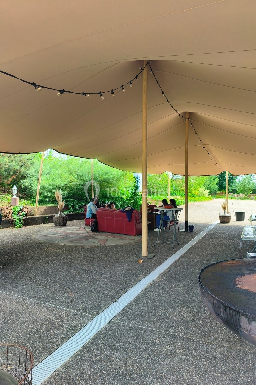 Espace extérieur couvert par une tente beige, avec des tables et des personnes assises dans un cadre verdoyant.