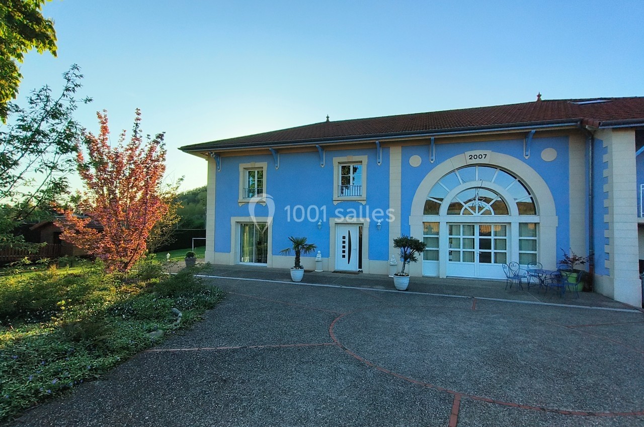 Façade d'une maison bleue avec une grande porte vitrée, entourée de plantes en pot et d'un arbre fleuri.