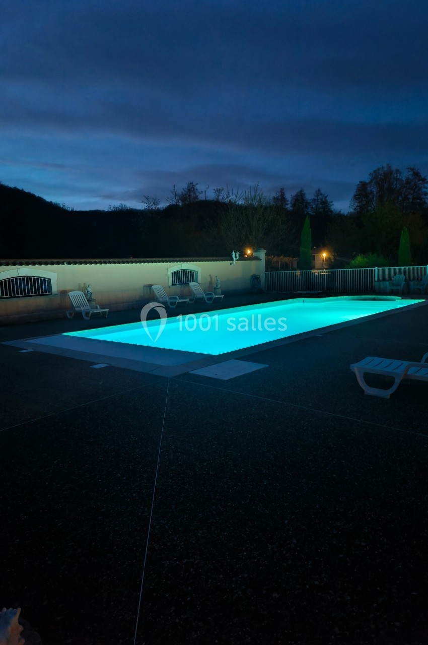 Piscine extérieure éclairée de nuit, entourée de chaises longues et d'arbres en arrière-plan.