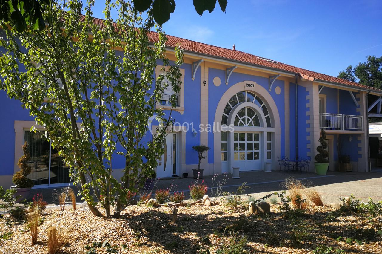 Façade d'un bâtiment bleu avec une grande porte vitrée en arc, entourée de végétation et d'un ciel dégagé.