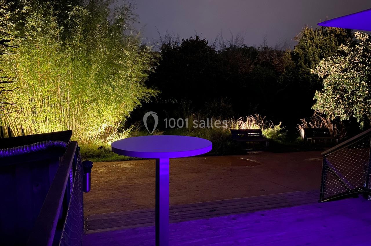 Terrasse en bois éclairée par des lumières violettes, avec une table haute et un jardin sombre en arrière-plan.