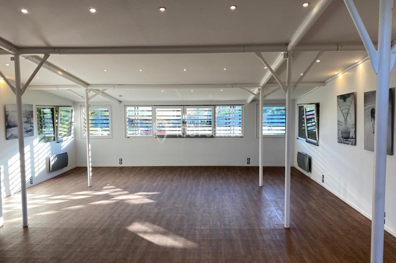 Salle lumineuse avec parquet en bois, murs blancs, fenêtres à persiennes et spots encastrés au plafond.