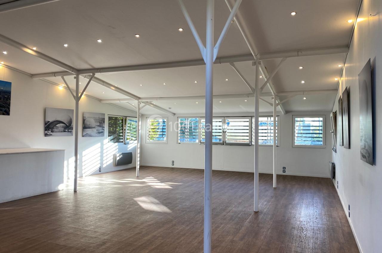 Salle lumineuse avec parquet, murs blancs, grandes fenêtres à persiennes et éclairage encastré au plafond.