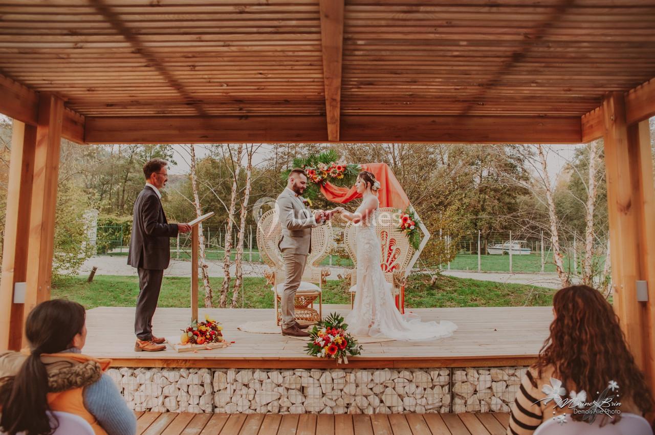 Un couple échange ses vœux de mariage sous une pergola en bois, entouré de fleurs et d'invités.