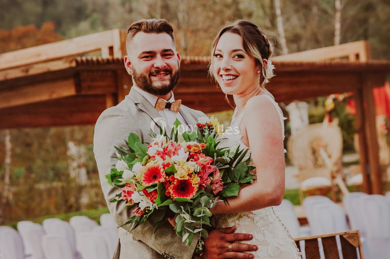 Un couple souriant en tenue de mariage pose devant une décoration extérieure avec des fleurs et des chaises blanches.