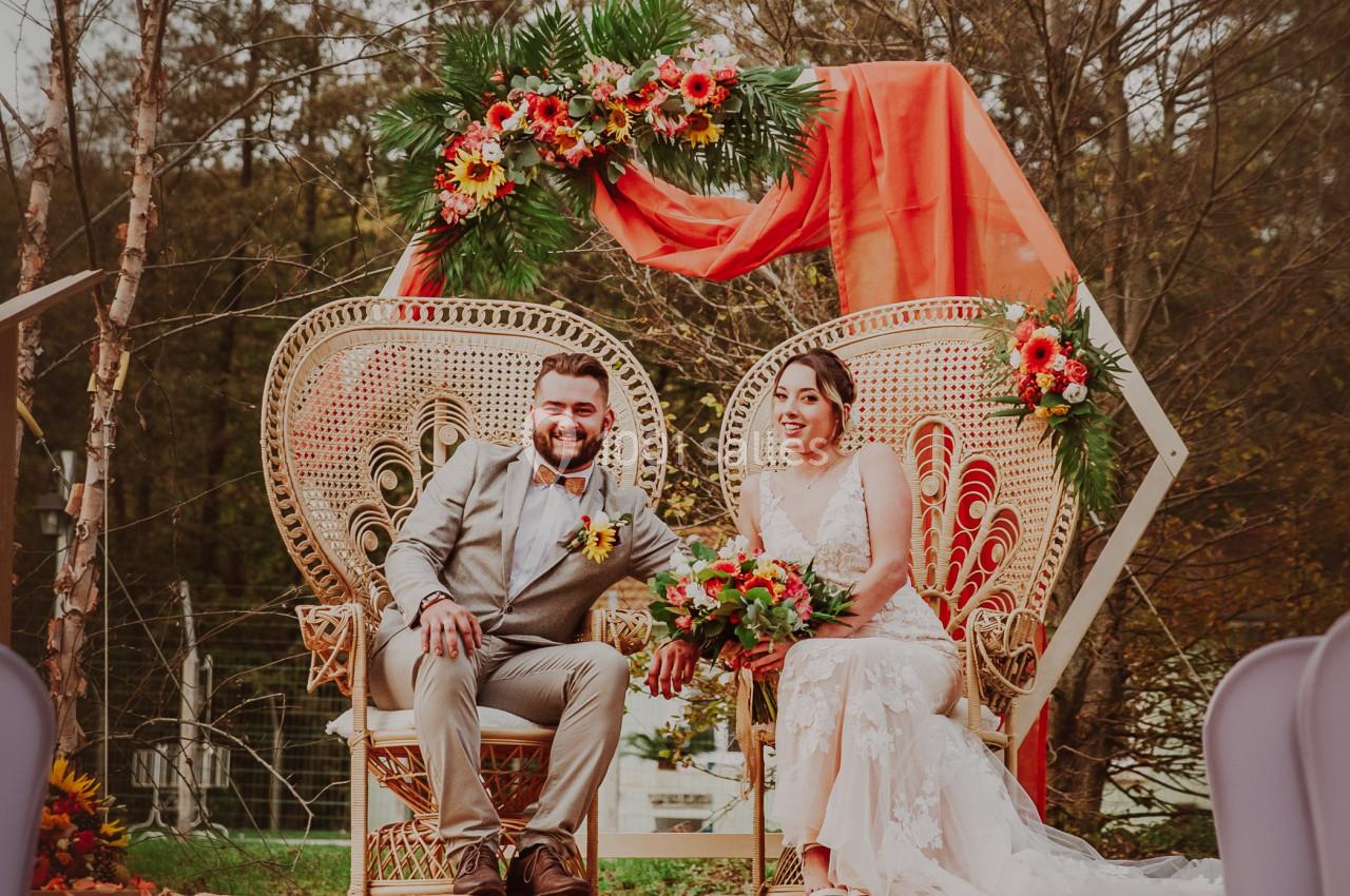 Un couple assis sur des fauteuils en osier décorés de fleurs, sous une pergola ornée de tissus et compositions florales.