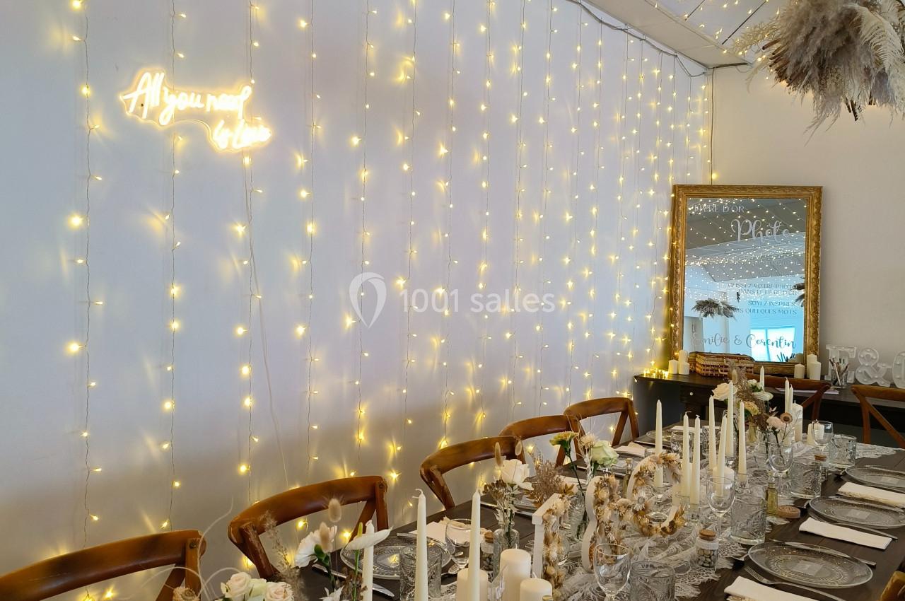 Table décorée pour un événement avec bougies, fleurs, vaisselle élégante et guirlandes lumineuses sur un mur blanc.