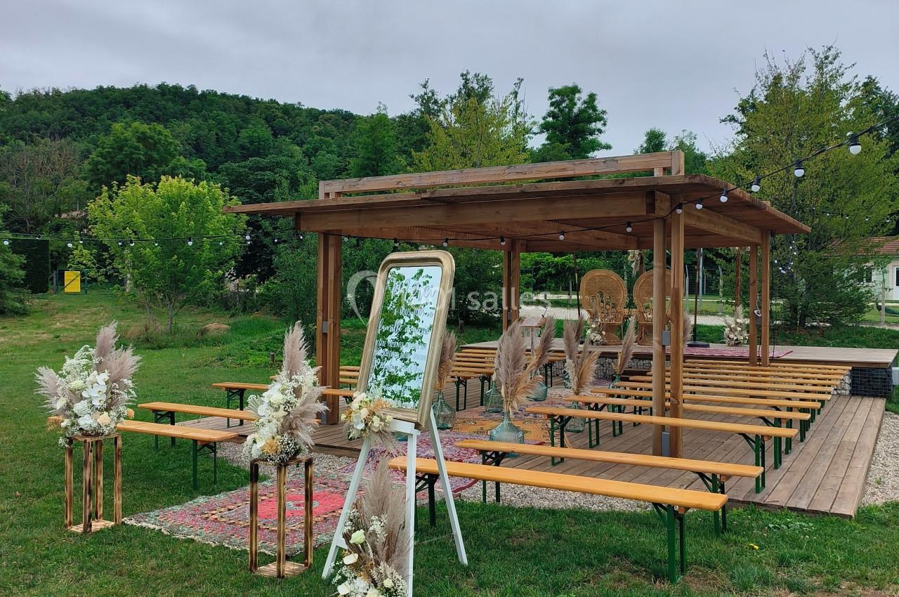 Espace extérieur aménagé pour une cérémonie, avec pergola en bois, bancs, décorations florales et miroir.