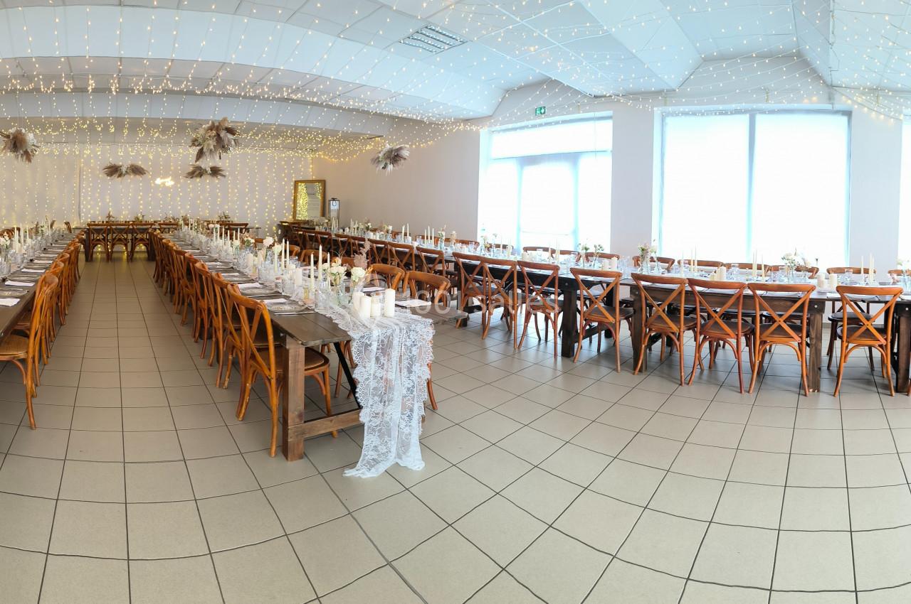 Salle de réception décorée avec des guirlandes lumineuses, tables en bois alignées et nappes en dentelle blanche.