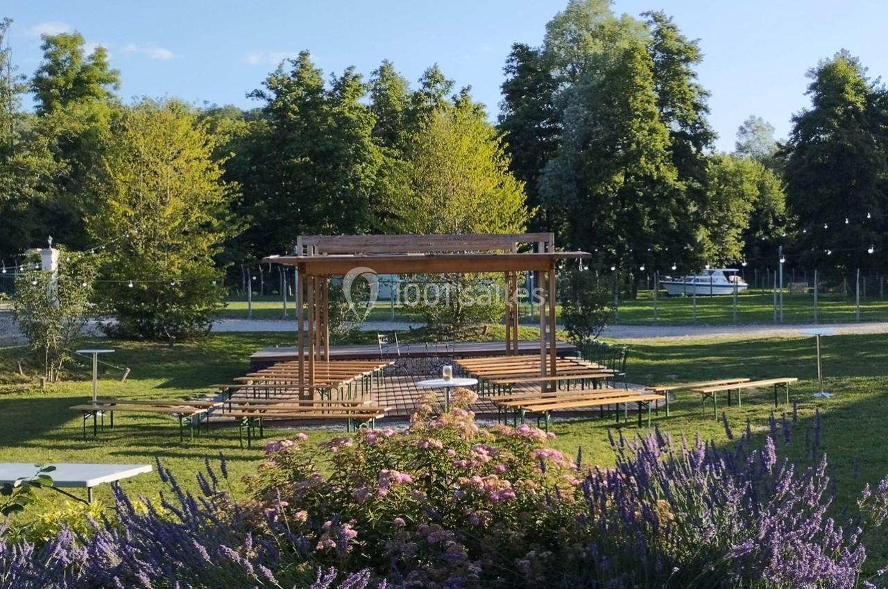 Structure en bois avec bancs installée dans un parc verdoyant, entourée de fleurs et d'arbres sous un ciel dégagé.