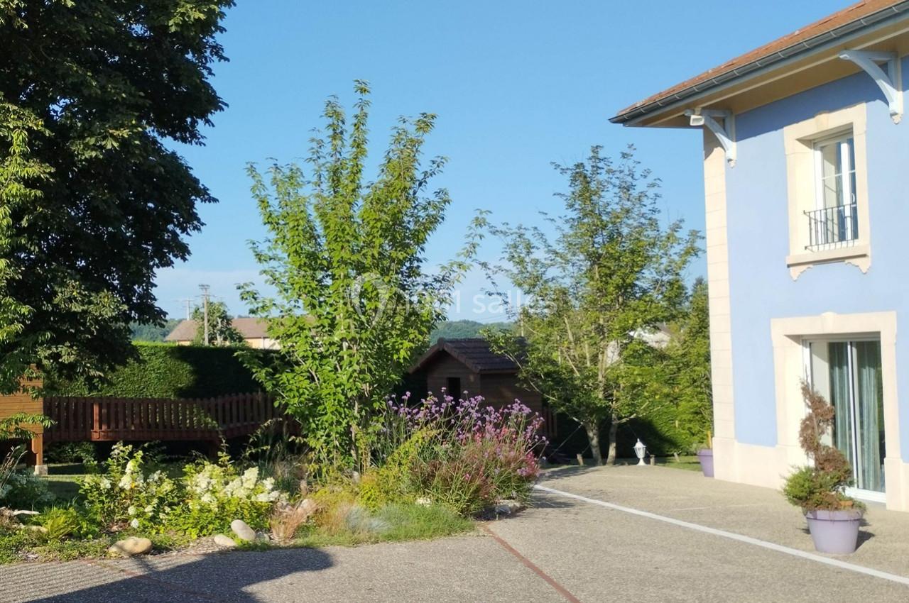 Allée pavée bordée de fleurs et d'arbustes, menant à une maison bleue et un jardin avec cabanon en arrière-plan.