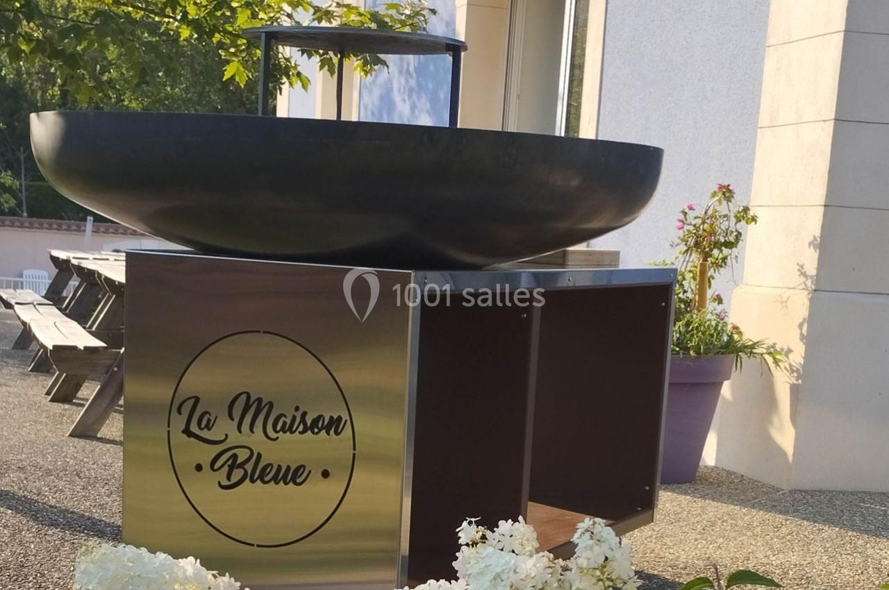 Brasero noir sur socle métallique marqué ’La Maison Bleue’, installé sur une terrasse ensoleillée avec fleurs et tables.