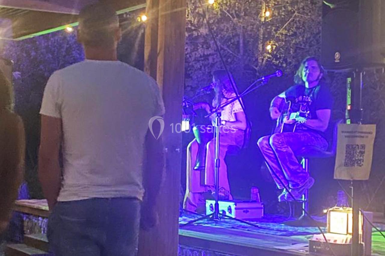 Un duo de musiciens joue de la guitare et chante sur une petite scène éclairée, devant un public en soirée.