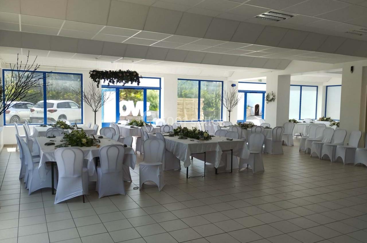 Salle lumineuse aménagée pour un événement, avec des tables décorées de nappes blanches et des chaises assorties.
