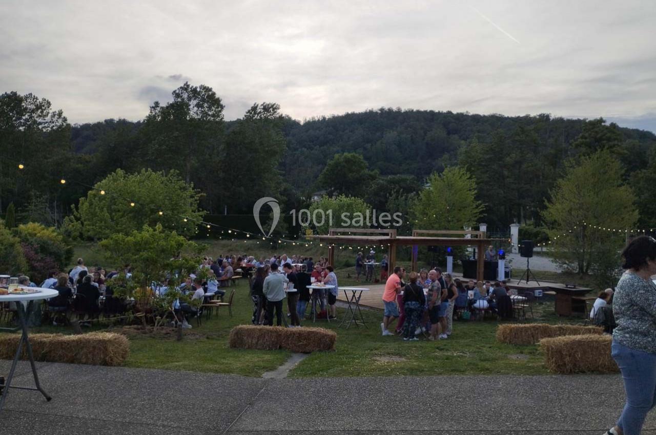 Personnes réunies en plein air autour de tables, avec guirlandes lumineuses et collines boisées en arrière-plan.
