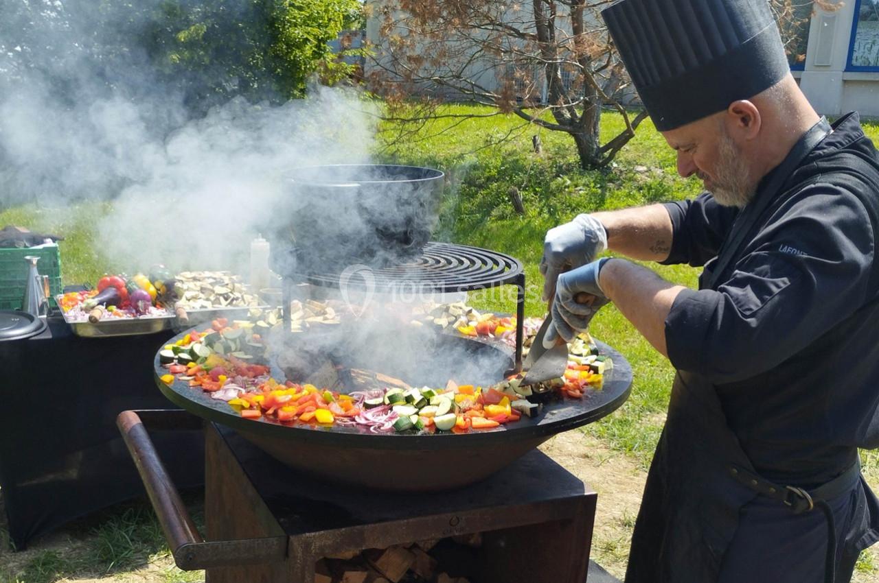 Un chef cuisine des légumes colorés sur une grande plancha en plein air, entouré de fumée et d'ustensiles.
