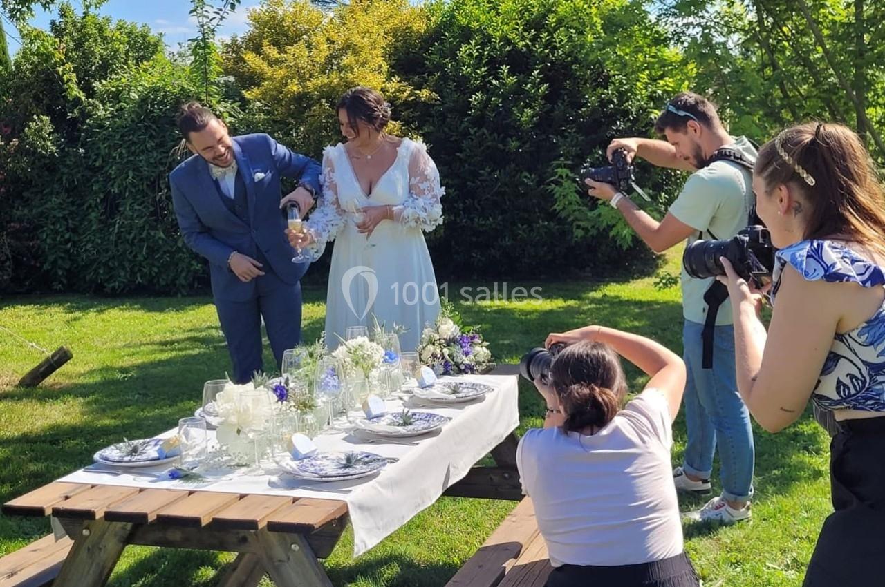 Un couple en tenue de mariage verse du champagne près d'une table décorée, entouré de photographes en extérieur.