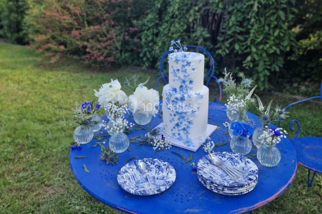 Gâteau décoré de fleurs bleues et blanches sur une table bleue avec vaisselle assortie, dans un jardin verdoyant.