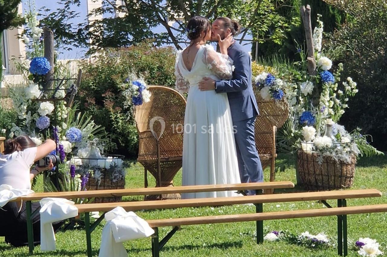 Un couple s'embrasse lors d'une cérémonie de mariage en extérieur, entouré de fleurs et de bancs décorés.