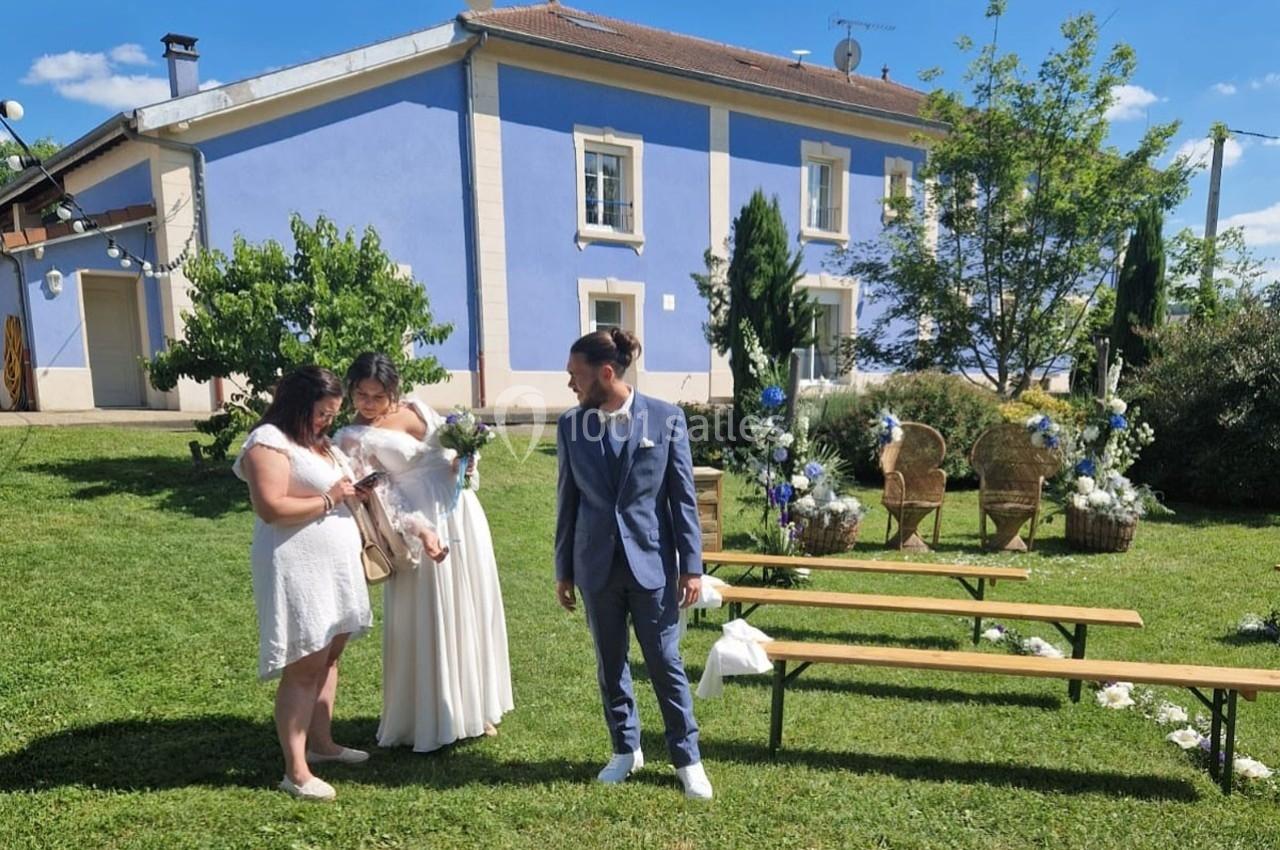 Un couple en tenue de mariage et une femme discutent dans un jardin devant une maison bleue par une journée ensoleillée.