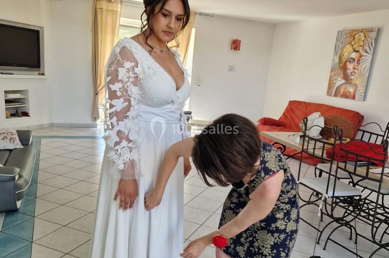 Une femme ajuste la robe blanche d'une mariée dans un salon lumineux avec des meubles modernes.