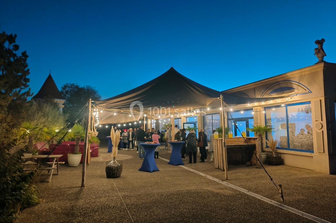 Terrasse éclairée par des guirlandes lumineuses sous une tente, avec des invités debout autour de tables hautes.