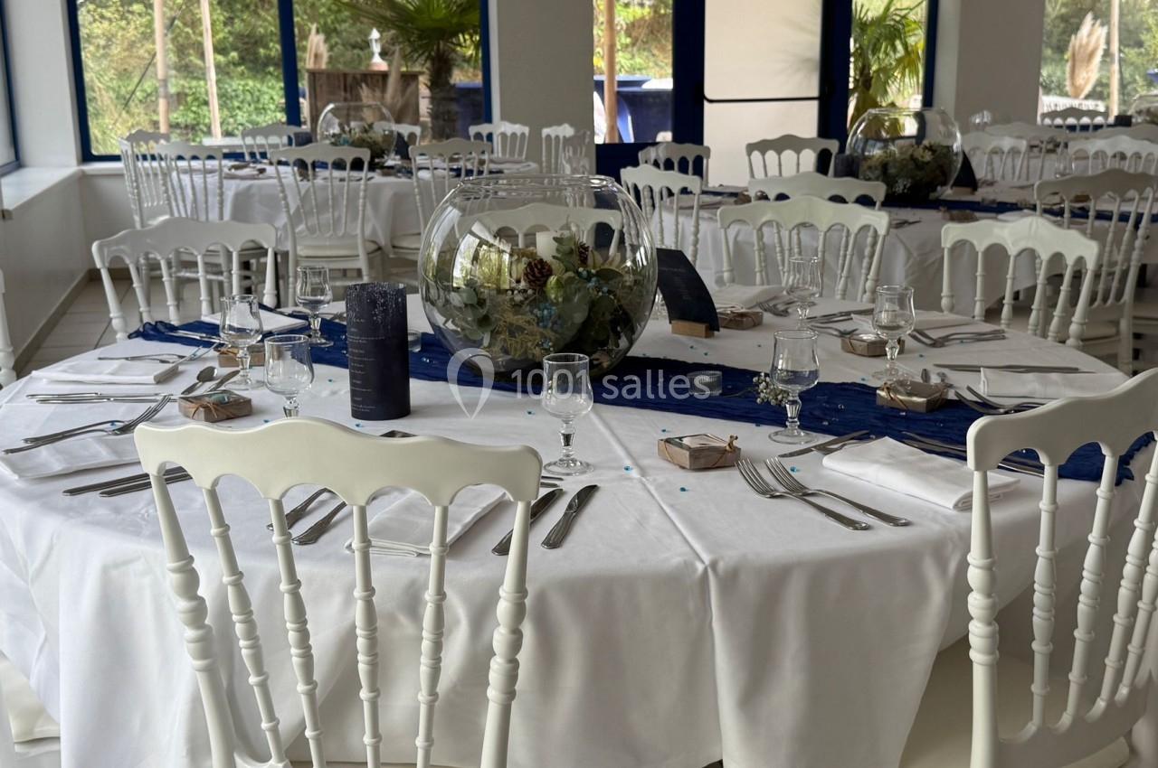 Salle de réception avec tables rondes dressées, nappes blanches, chaises blanches et centres de table décoratifs.