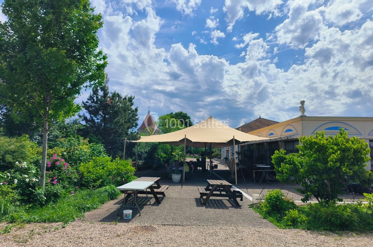 Espace extérieur avec tables de pique-nique sous des voiles d'ombrage, entouré de verdure et d'un ciel partiellement nuageux.
