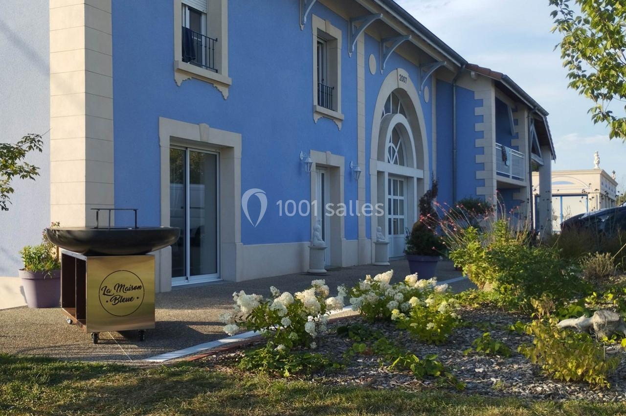 Façade d'un bâtiment bleu avec des fenêtres, un parterre fleuri et un panneau indiquant ’La Maison Rose’.