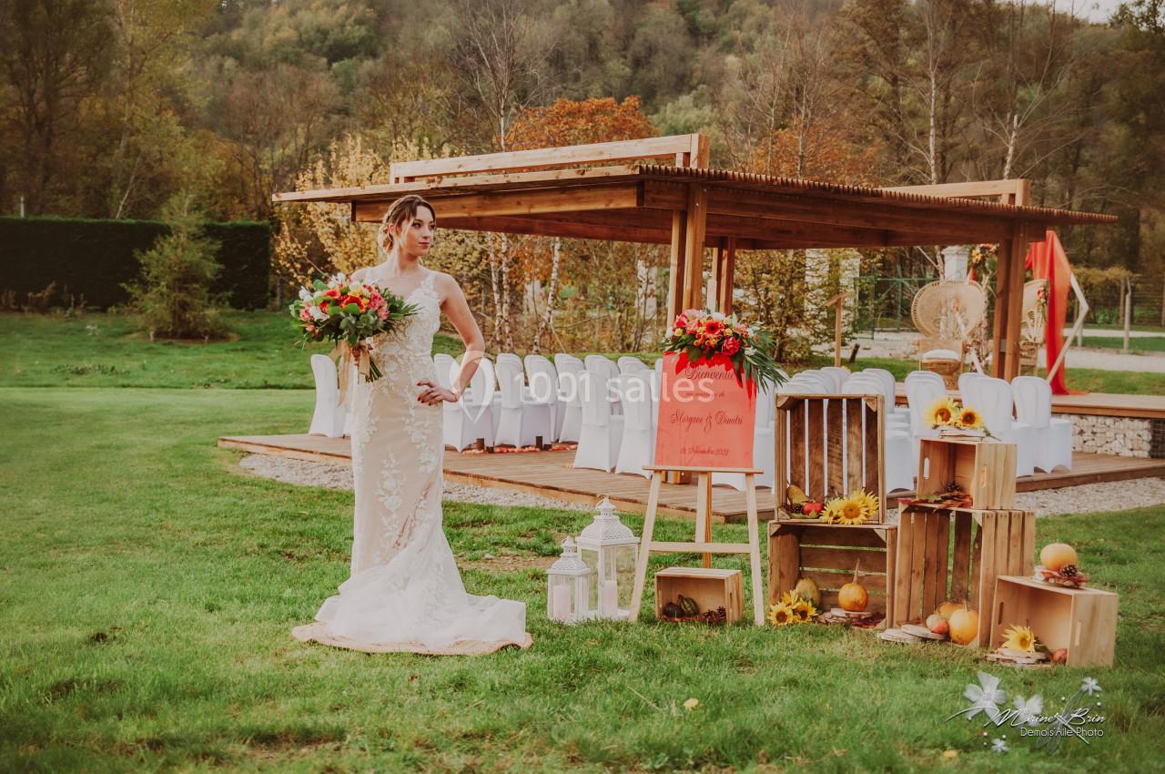 Une mariée en robe blanche tient un bouquet devant une cérémonie en extérieur, décorée de fleurs et de citrouilles.