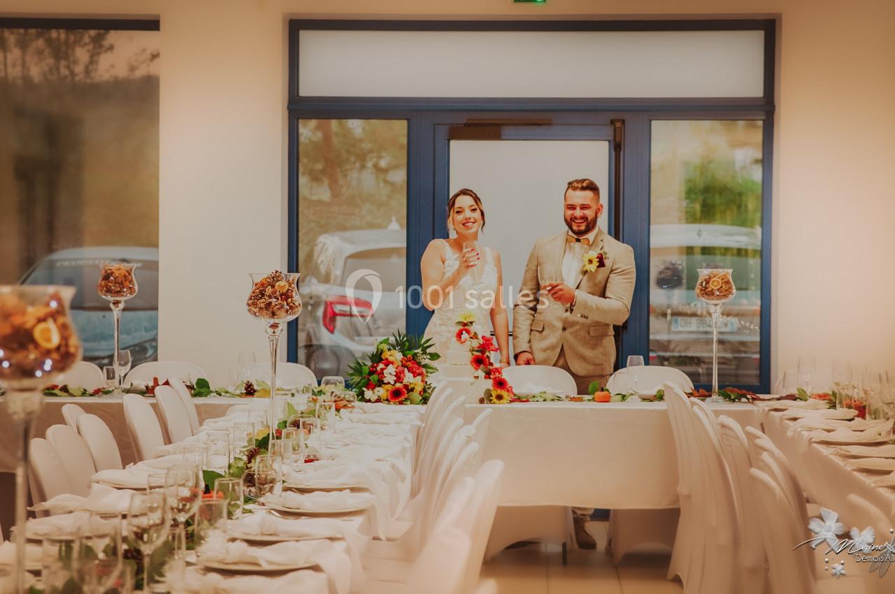 Un couple en tenue de mariage sourit debout derrière une table décorée dans une salle de réception.