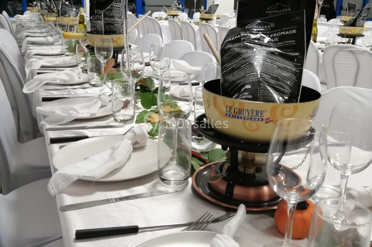 Tables dressées pour un repas, avec nappes blanches, assiettes, verres, décorations et un caquelon à fondue au premier plan.