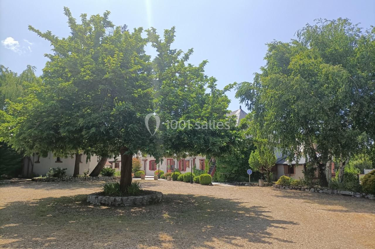 Arbre au centre d'une cour en gravier, entouré de bâtiments avec volets rouges et végétation sous un ciel dégagé.