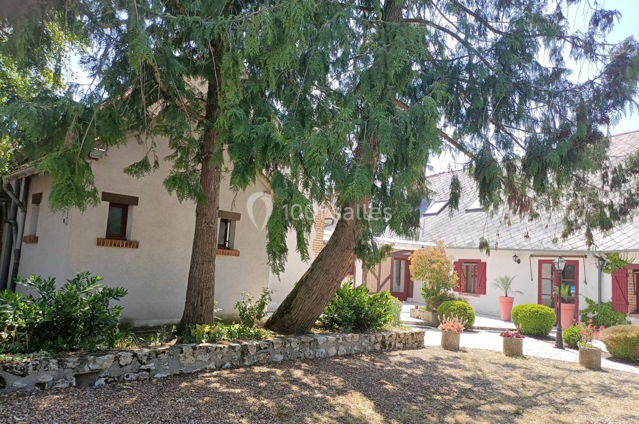 Cour d'une maison avec façade blanche, volets rouges, arbres imposants et jardinières fleuries sous un ciel dégagé.