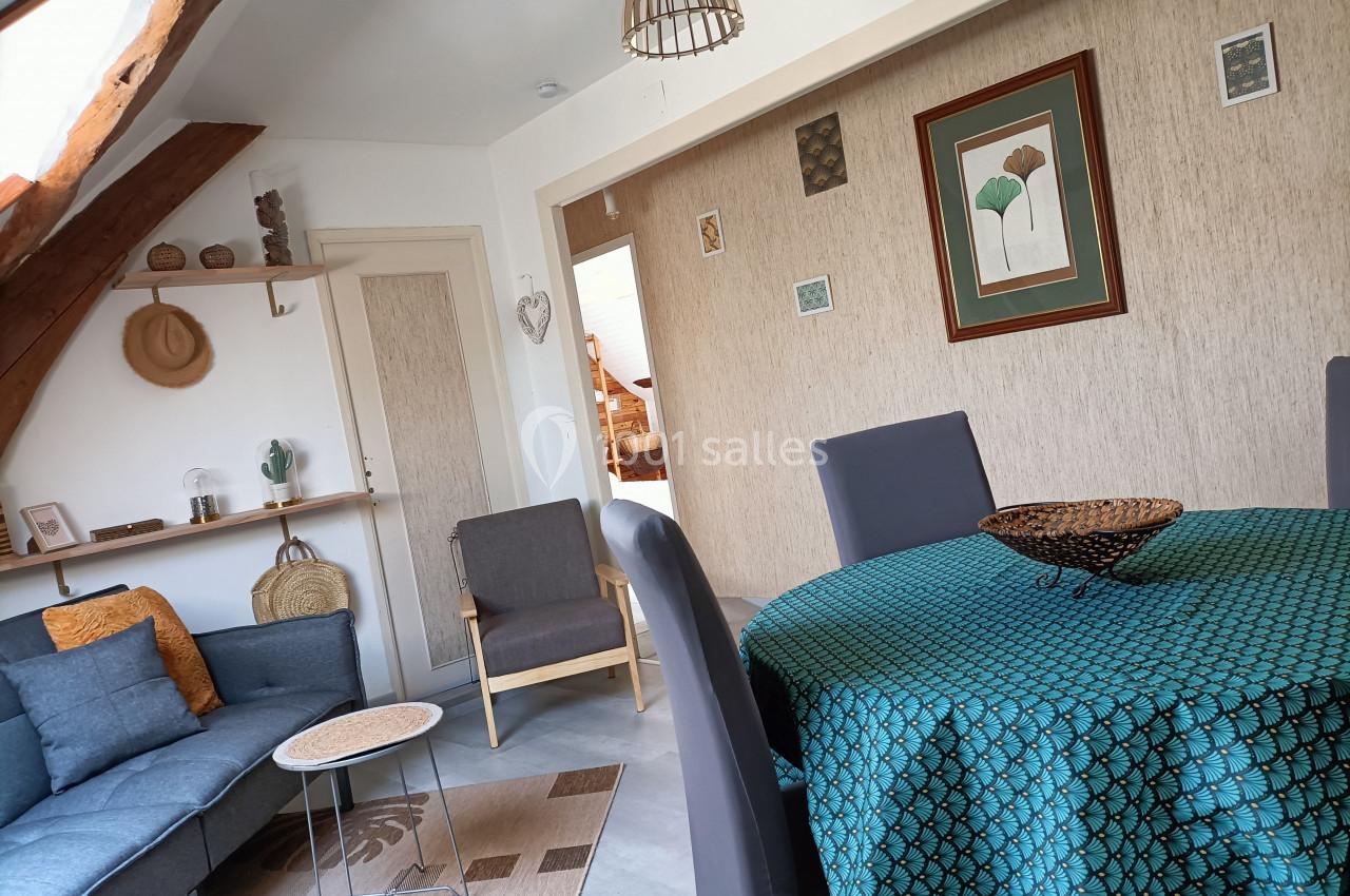 Salon lumineux avec poutres apparentes, table ronde habillée d'une nappe verte, canapé gris et décorations murales.