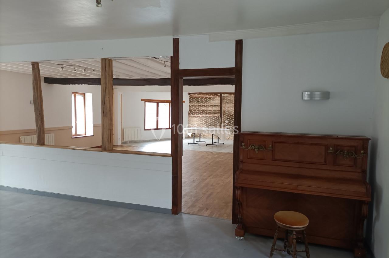 Salle lumineuse avec parquet, murs blancs, poutres apparentes, un piano en bois et un tabouret au premier plan.