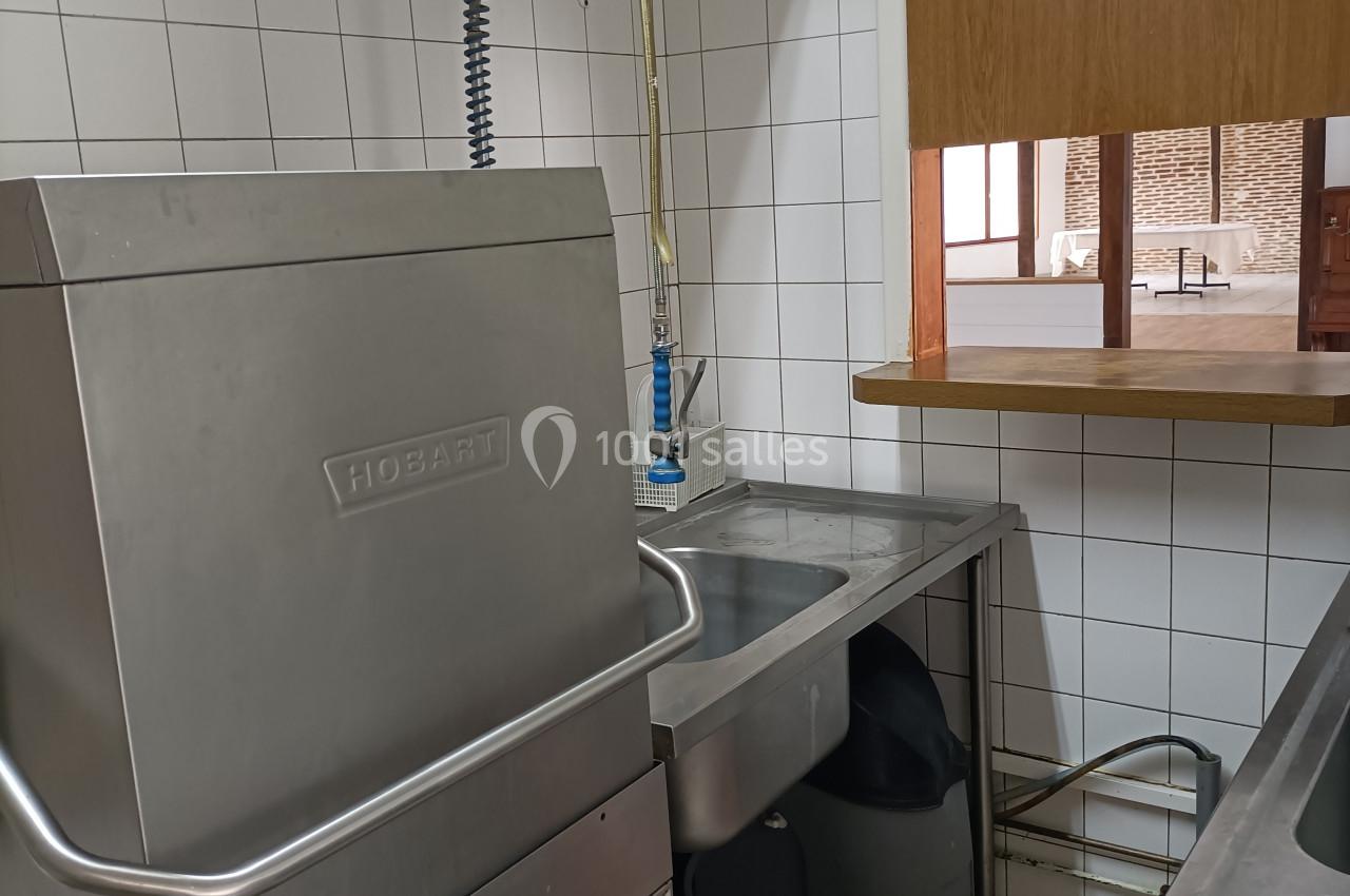 Lave-vaisselle industriel en inox dans une cuisine carrelée blanche, avec un évier et un robinet flexible à proximité.