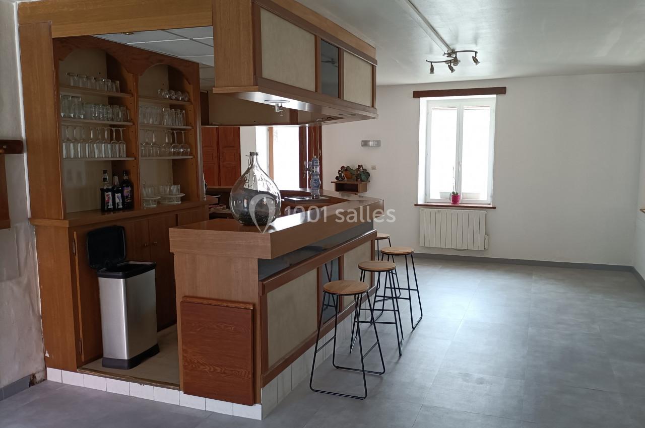 Cuisine ouverte avec un bar en bois, tabourets hauts, étagères avec verres et bouteilles, et lumière naturelle.