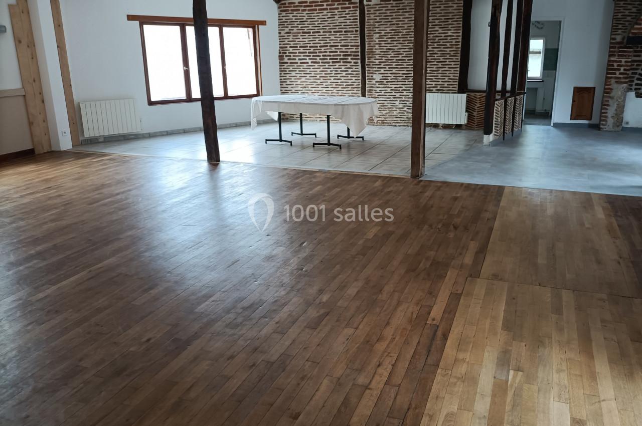 Salle vide avec parquet en bois, murs en briques et fenêtres laissant entrer la lumière naturelle.